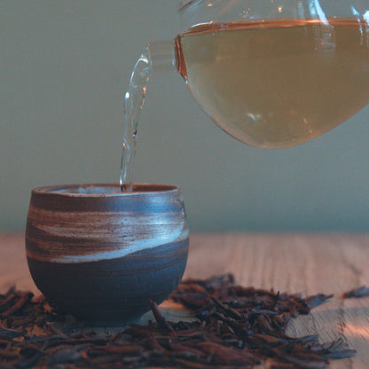 Wild purple raw puerh tea being poured into a small cup with loose leaf tea on the table surrounding it. 